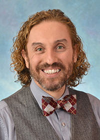 Portrait of Edward Moreira Bahnson. Edward is a white man with curly brown and blond hair and a brown and gray beard. He is wearing a striped shirt, gray wool vest and red plaid bowtie and smiling at the camera.